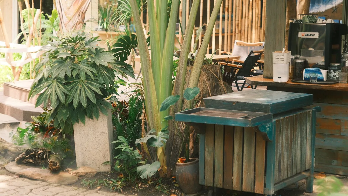 Outdoor cafe setting with lush greenery, a rustic wooden cart, and chairs.